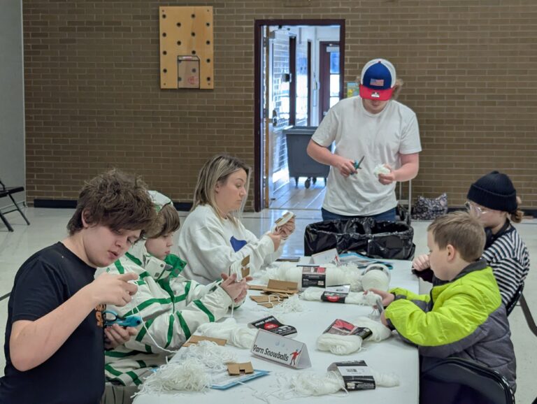 A group of students and teacher sit at a table making yarn snowballs.